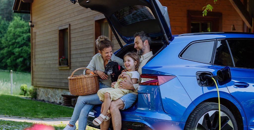 Familie sitzt entspannt am Kofferraum, während ihr E-Auto zu Hause lädt