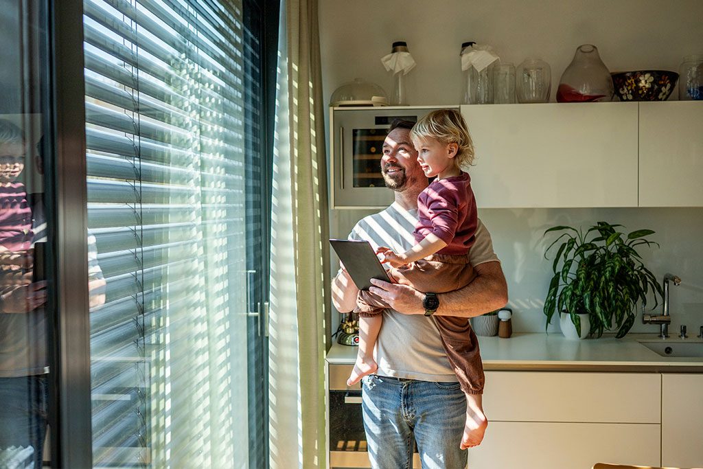 Vater mit Kind am Fenster, die Rollläden öffnen sich mit dem Sonnenaufgang